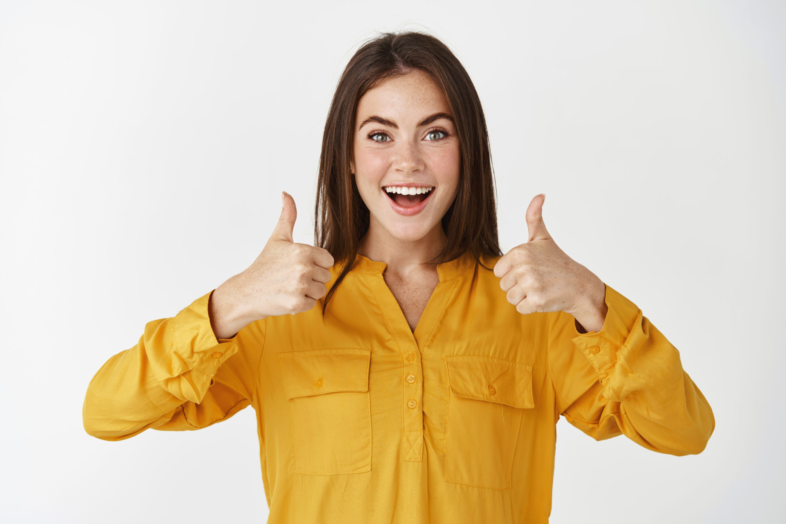 Impressed young woman showing thumbs up and smiling amazed, praising something cool, standing over white background.